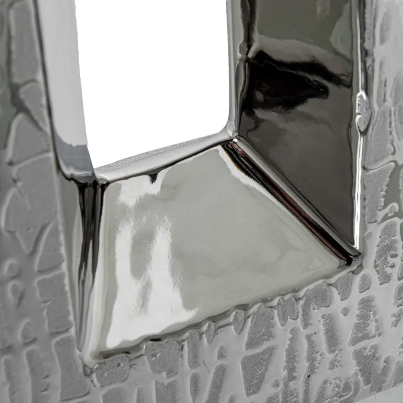 Vaso a bottiglia rettangolare verticale firmato Camacho, caratterizzato da un'elegante finitura ceramica color argento lucido con una texture a rilievo finemente lavorata e un originale design geometrico irregolare con foro centrale vuoto. Questo moderno complemento d'arredo è provvisto di un collo stretto superiore ideale per ospitare singoli steli floreali, perfetto come sofisticato oggetto ornamentale per impreziosire mobili, consolle o tavoli in contesti contemporanei.