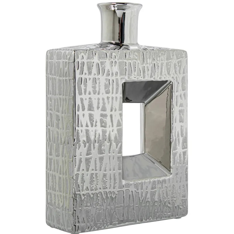 Vaso a bottiglia rettangolare verticale firmato Camacho, caratterizzato da un'elegante finitura ceramica color argento lucido con una texture a rilievo finemente lavorata e un originale design geometrico irregolare con foro centrale vuoto. Questo moderno complemento d'arredo è provvisto di un collo stretto superiore ideale per ospitare singoli steli floreali, perfetto come sofisticato oggetto ornamentale per impreziosire mobili, consolle o tavoli in contesti contemporanei.