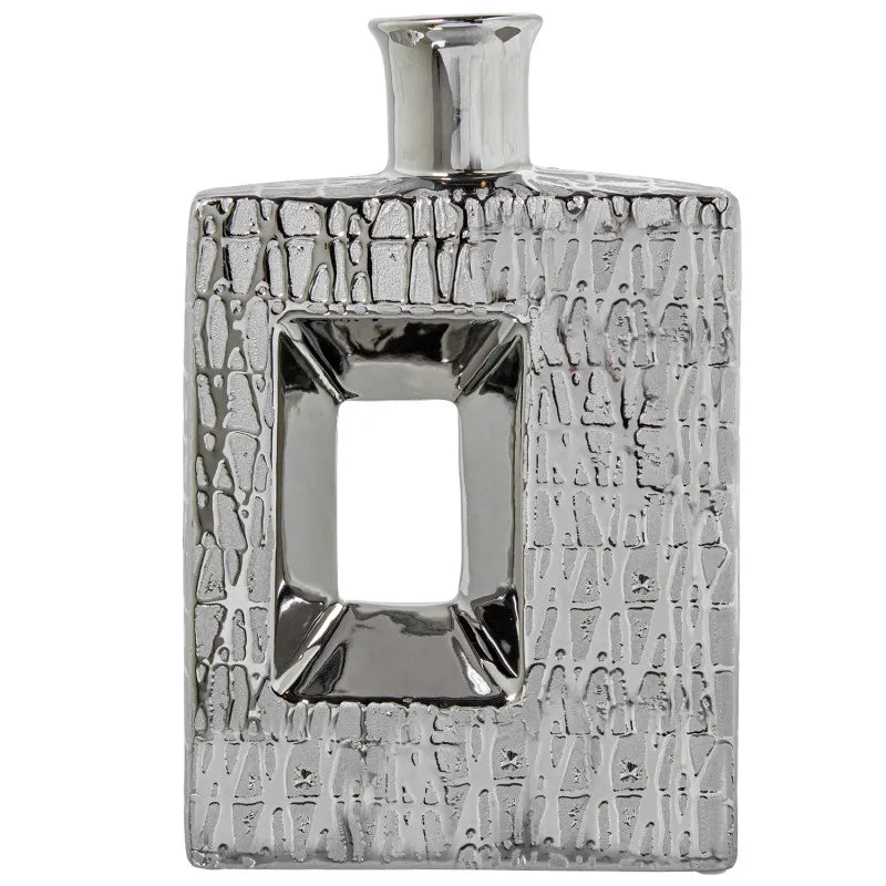 Vaso a bottiglia rettangolare verticale firmato Camacho, caratterizzato da un'elegante finitura ceramica color argento lucido con una texture a rilievo finemente lavorata e un originale design geometrico irregolare con foro centrale vuoto. Questo moderno complemento d'arredo è provvisto di un collo stretto superiore ideale per ospitare singoli steli floreali, perfetto come sofisticato oggetto ornamentale per impreziosire mobili, consolle o tavoli in contesti contemporanei.