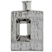 Vaso a bottiglia rettangolare verticale firmato Camacho, caratterizzato da un'elegante finitura ceramica color argento lucido con una texture a rilievo finemente lavorata e un originale design geometrico irregolare con foro centrale vuoto. Questo moderno complemento d'arredo è provvisto di un collo stretto superiore ideale per ospitare singoli steli floreali, perfetto come sofisticato oggetto ornamentale per impreziosire mobili, consolle o tavoli in contesti contemporanei.