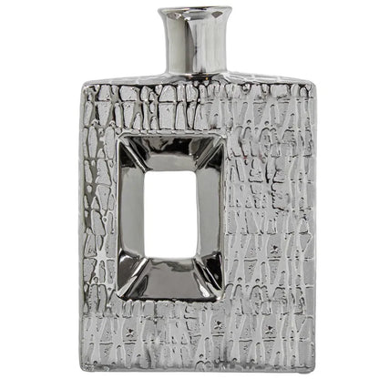 Vaso a bottiglia rettangolare verticale firmato Camacho, caratterizzato da un'elegante finitura ceramica color argento lucido con una texture a rilievo finemente lavorata e un originale design geometrico irregolare con foro centrale vuoto. Questo moderno complemento d'arredo è provvisto di un collo stretto superiore ideale per ospitare singoli steli floreali, perfetto come sofisticato oggetto ornamentale per impreziosire mobili, consolle o tavoli in contesti contemporanei.