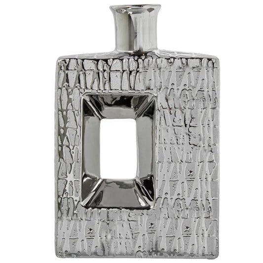 Vaso a bottiglia rettangolare verticale firmato Camacho, caratterizzato da un'elegante finitura ceramica color argento lucido con una texture a rilievo finemente lavorata e un originale design geometrico irregolare con foro centrale vuoto. Questo moderno complemento d'arredo è provvisto di un collo stretto superiore ideale per ospitare singoli steli floreali, perfetto come sofisticato oggetto ornamentale per impreziosire mobili, consolle o tavoli in contesti contemporanei.