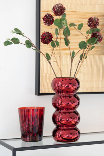 Vaso in vetro rosso scuro modello Bruco firmato Jolipa, caratterizzato da un design moderno e sinuoso a sezioni sferiche sovrapposte con finitura lucida e metallizzata. Questo originale vaso decorativo di design è perfetto come centrotavola o portafiori elegante per arredare con uno stile ricercato e contemporaneo gli interni della casa.