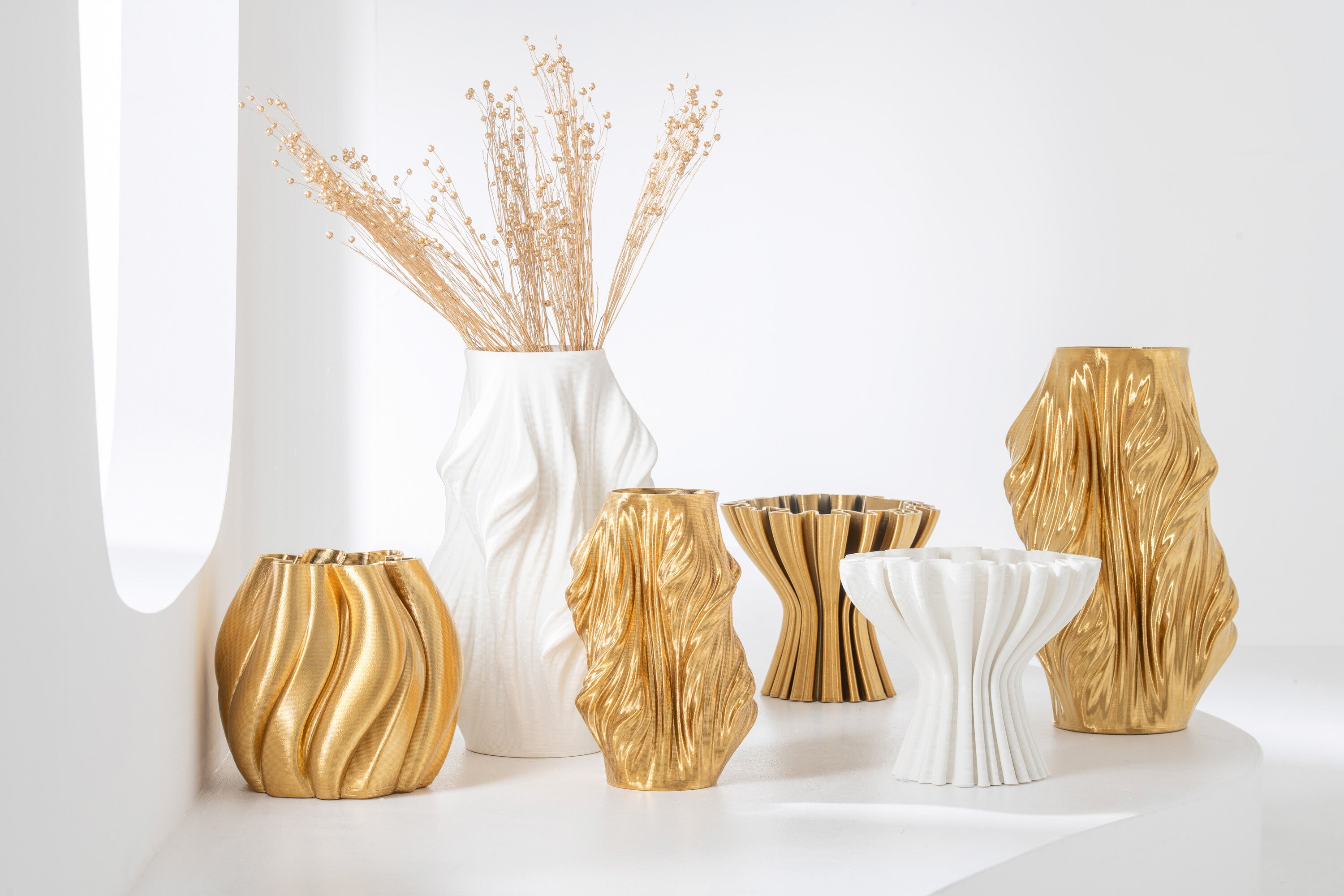 Composizione di vasi in porcellana della collezione 3D in vari formati, colori oro e bianco opaco. I vasi presentano forme scultoree asimmetriche e texture rigate, disposti su un piano bianco minimalista.