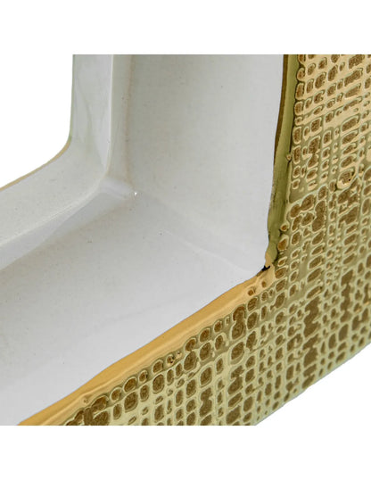 Vaso a bottiglia rettangolare orizzontale firmato Camacho, caratterizzato da un'elegante finitura ceramica color oro lucido con una texture a rilievo finemente lavorata, dettagli bianchi e un originale design geometrico con foro centrale vuoto. Questo moderno complemento d'arredo è provvisto di un collo stretto superiore ideale per ospitare singoli steli floreali, perfetto come sofisticato oggetto ornamentale per impreziosire mobili, consolle o tavoli in contesti contemporanei.