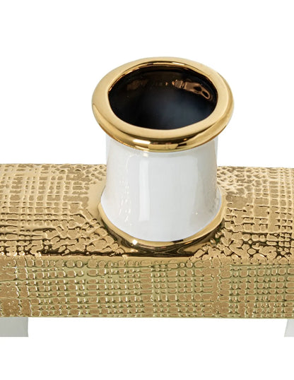 Vaso a bottiglia rettangolare orizzontale firmato Camacho, caratterizzato da un'elegante finitura ceramica color oro lucido con una texture a rilievo finemente lavorata, dettagli bianchi e un originale design geometrico con foro centrale vuoto. Questo moderno complemento d'arredo è provvisto di un collo stretto superiore ideale per ospitare singoli steli floreali, perfetto come sofisticato oggetto ornamentale per impreziosire mobili, consolle o tavoli in contesti contemporanei.
